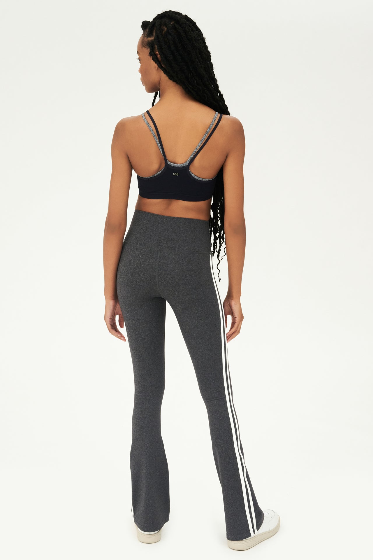 Full back view of woman with black braided hair wearing dark gray high waist below ankle length legging with wide flared bottoms and double white stripes on both legs. Also double dark gray and black racerback bra with spaghetti straps. Paired with white shoes.