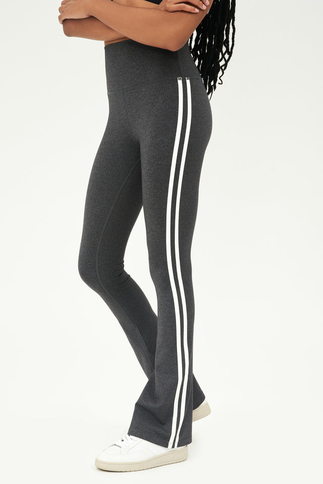 Front side view of woman wearing dark gray high waist below ankle length legging with wide flared bottoms and double white stripes on both legs. Paired with white shoes.