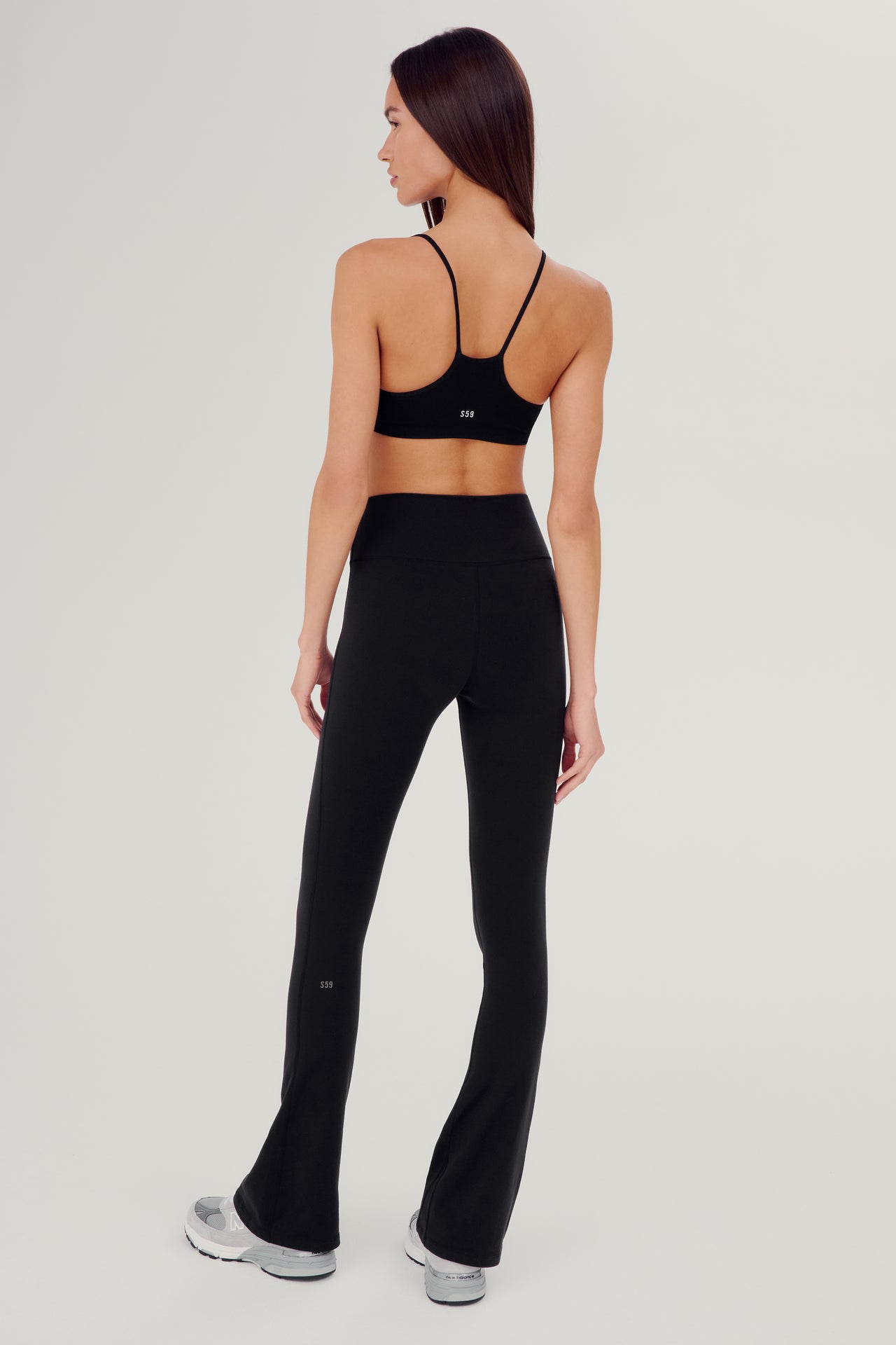 Full back view of woman wearing black high waist below ankle length legging with wide flared bottoms and gray S59 logo on bottom left calf. Also wearing black racerback bra with spaghetti straps and white S59 logo on back. Paired with white and gray shoes.