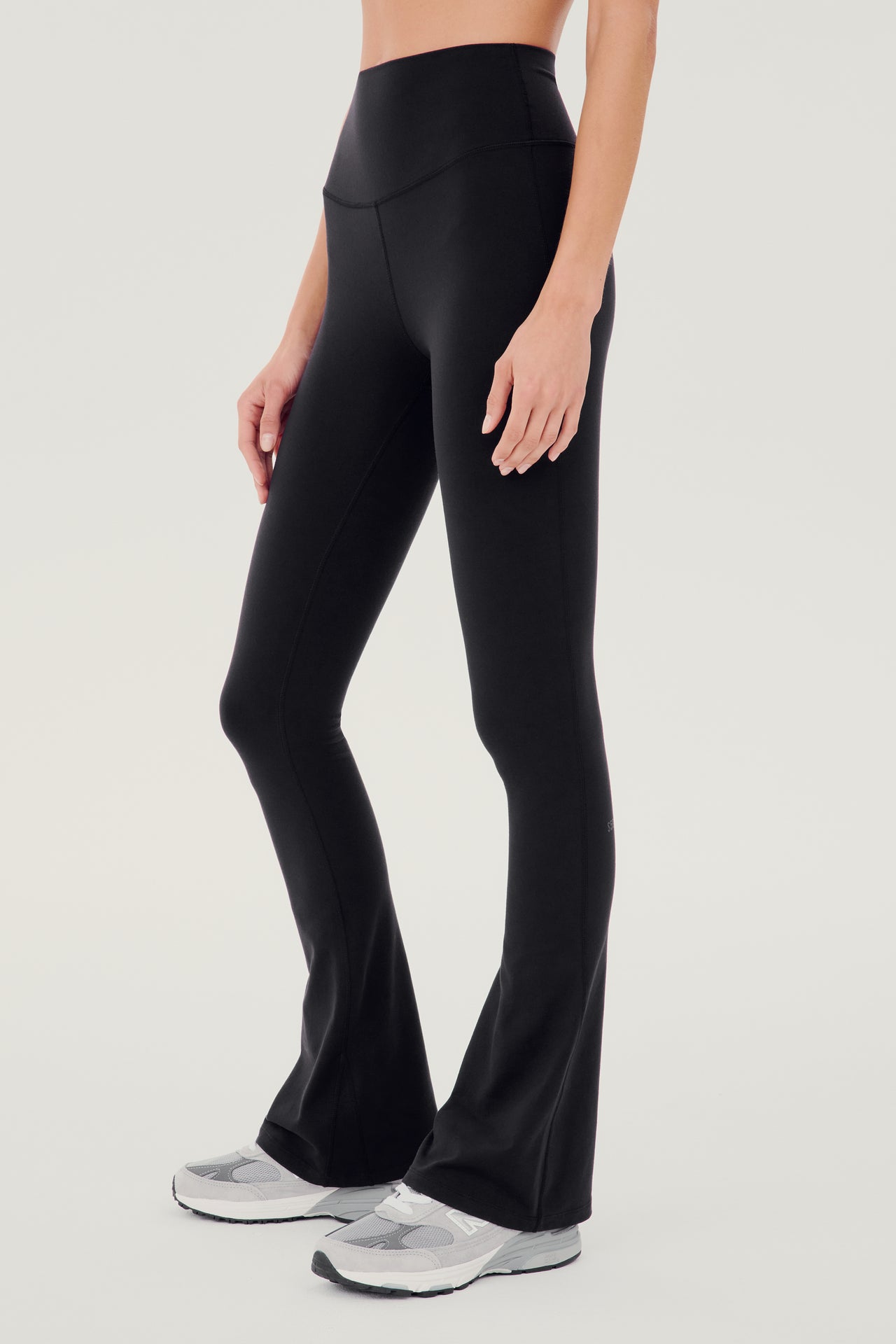 Front side view of woman wearing black high waist below ankle length legging with wide flared bottoms. Paired with white and gray shoes.