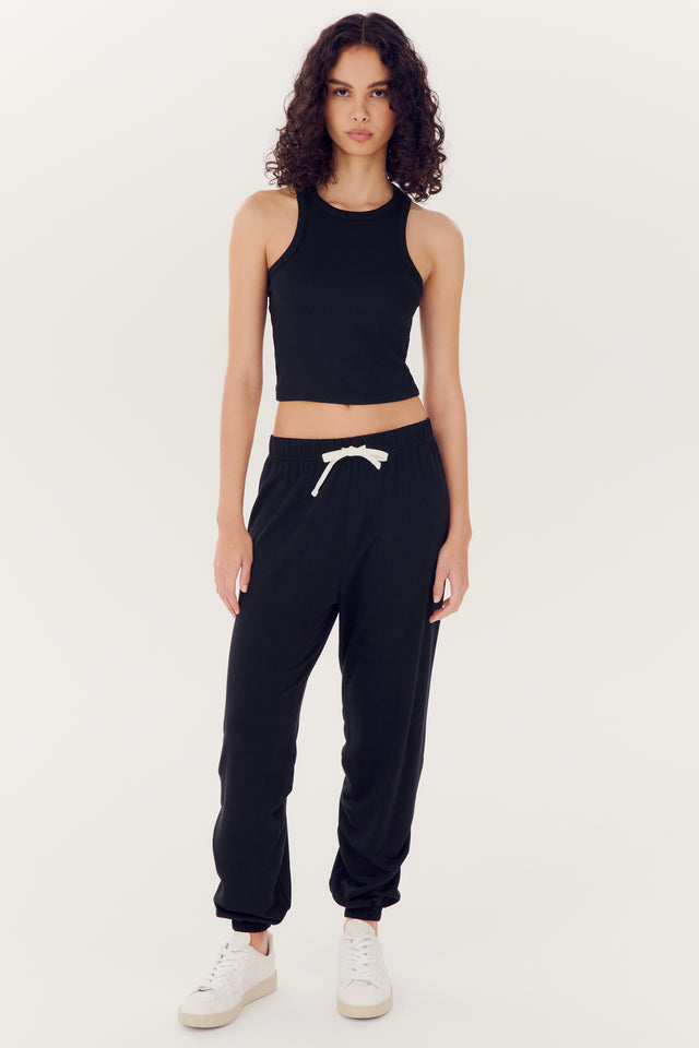 A woman wearing a comfortable black tank top made of modal fabric, Andie Oversized Fleece Sweatpant - Black trousers, and white sneakers stands against a plain background.