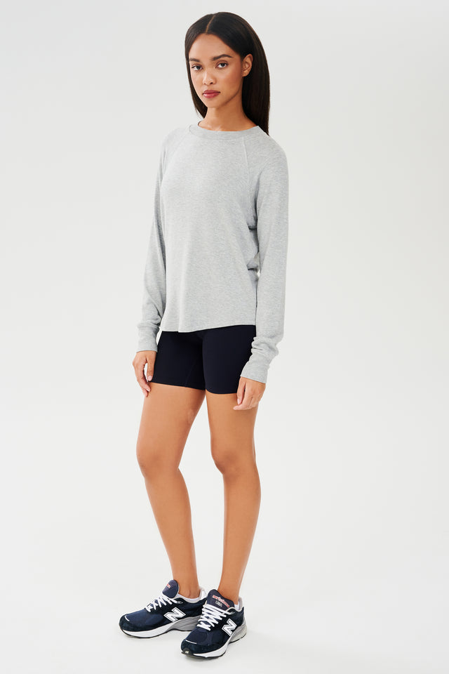 Full front side shot of woman with dark straight hair, wearing grey sweatshirt with black shorts and dark blue shoes
