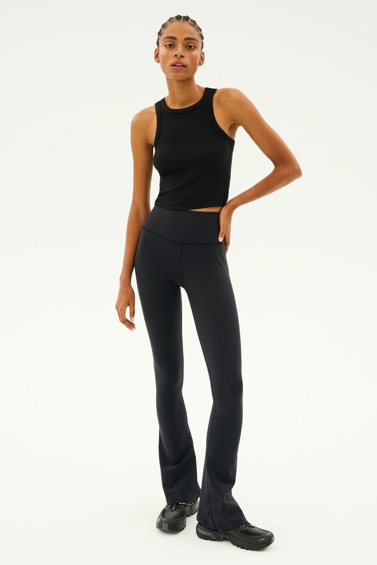 Person wearing the Kiki Rib Crop Tank in black by SPLITS59, with high-waisted black pants and black shoes, stands with one hand on hip against a plain white background.