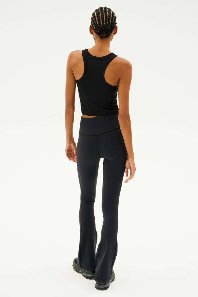 A person wearing the Kiki Rib Crop Tank in black by SPLITS59 and black flared leggings, walking away on a plain background.