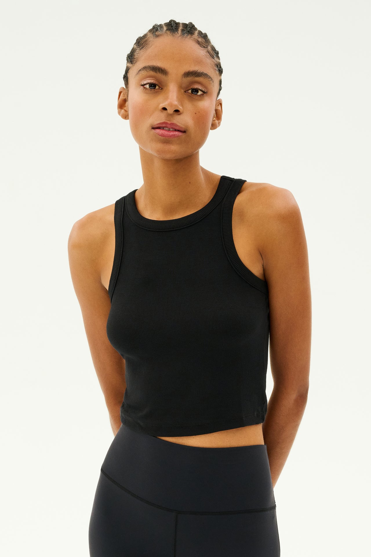 Wearing a Kiki Rib Crop Tank in black by SPLITS59, paired with leggings made from expertly crafted baby rib fabric, an individual stands confidently against a plain background.