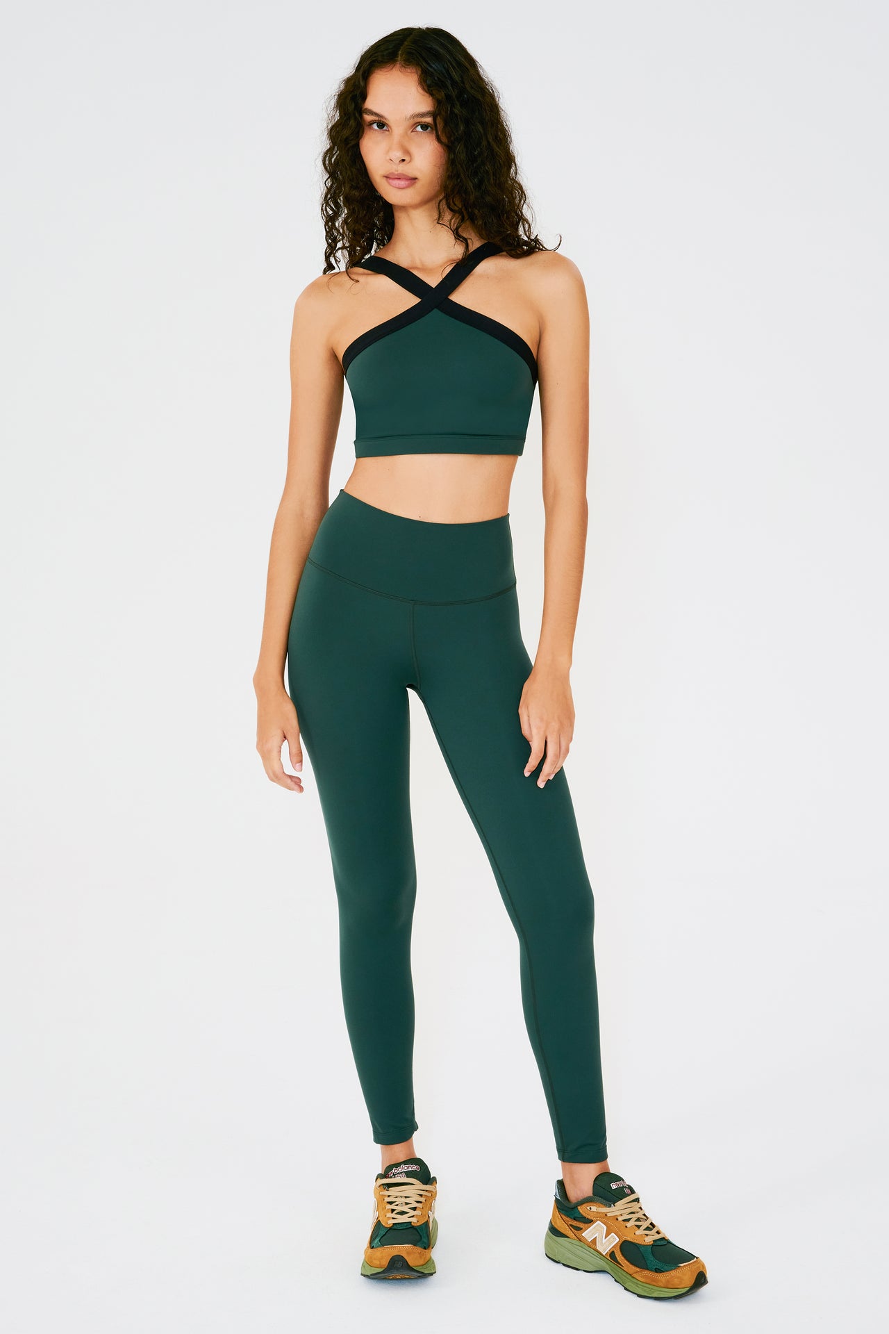 Front full view of woman with dark curly hair wearing dark green high waist  leggings and dark green bra with black crossed straps paired with dark green and orange shoes