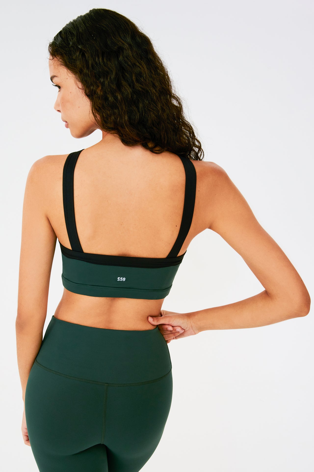 Back view of woman with dark wavy hair wearing a dark green bra with thick black straps and white S59 logo and dark green leggings