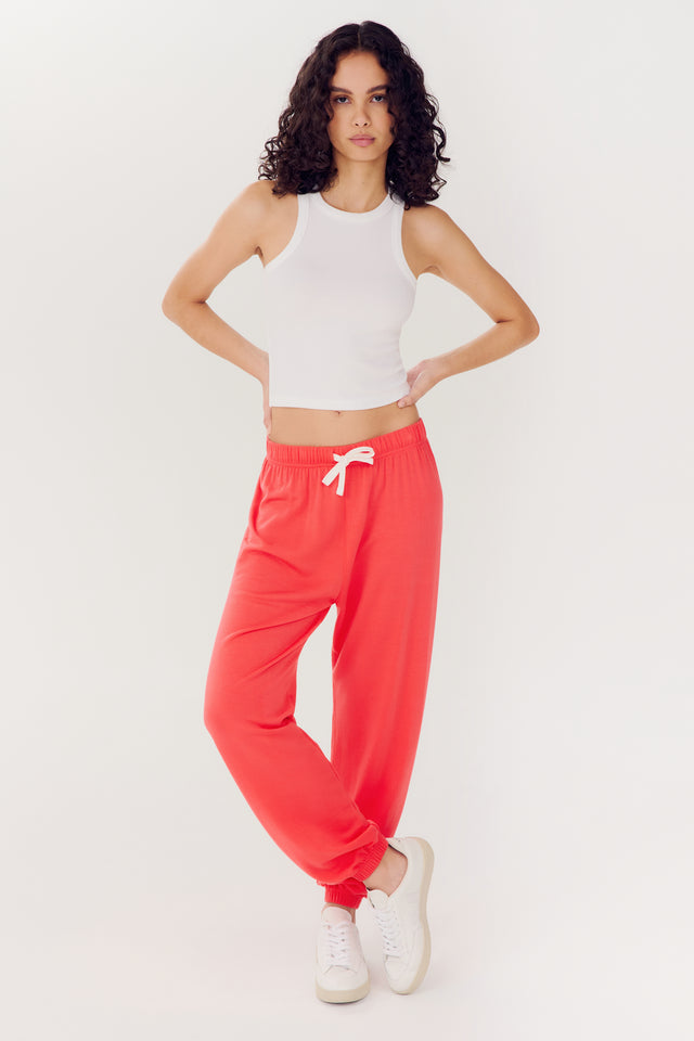 Woman posing in a white tank top and red SPLITS59 Andie Oversized Fleece Sweatpants - Melon with white sneakers against a plain background.