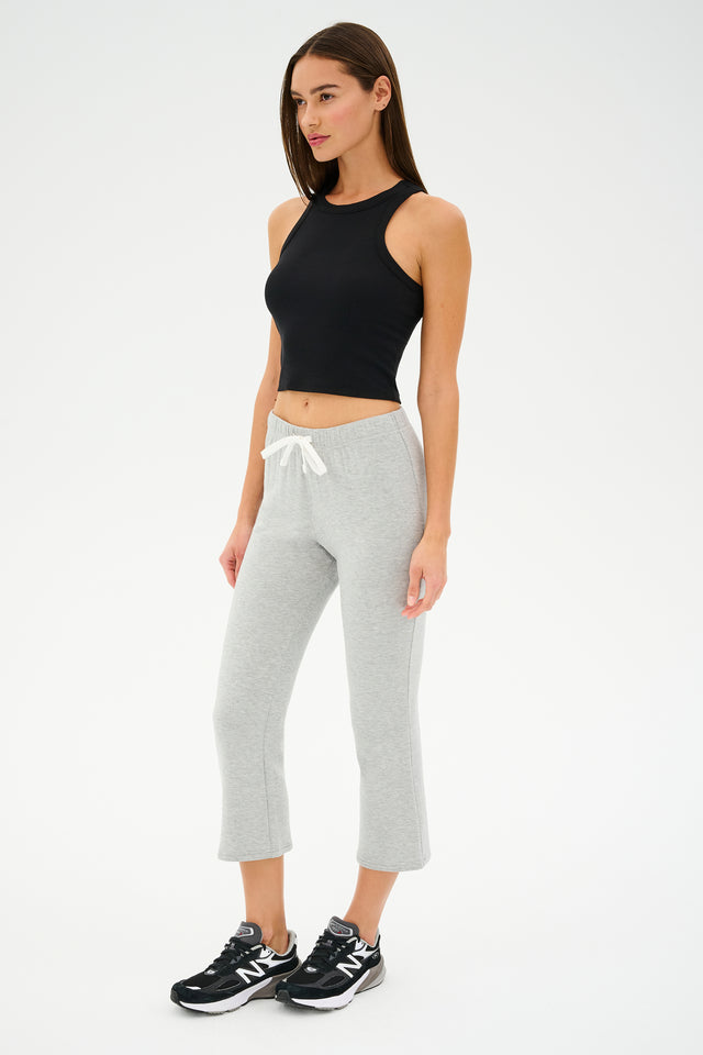 A woman in comfy casual chic stands against a white background, wearing a black sleeveless top with Brooks Fleece Cropped Flare pants in Heather Grey by SPLITS59 and sleek black New Balance sneakers.