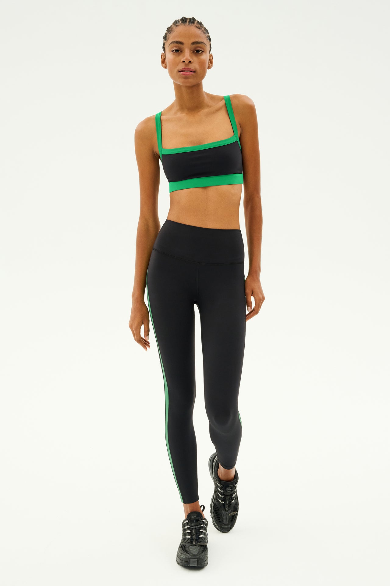 A person wearing the Monah Rigor Bra in Black/Grass from SPLITS59, paired with matching leggings, confidently strides forward on a white background, showcasing the sleek and flexible nylon-spandex fabric of this athletic outfit.