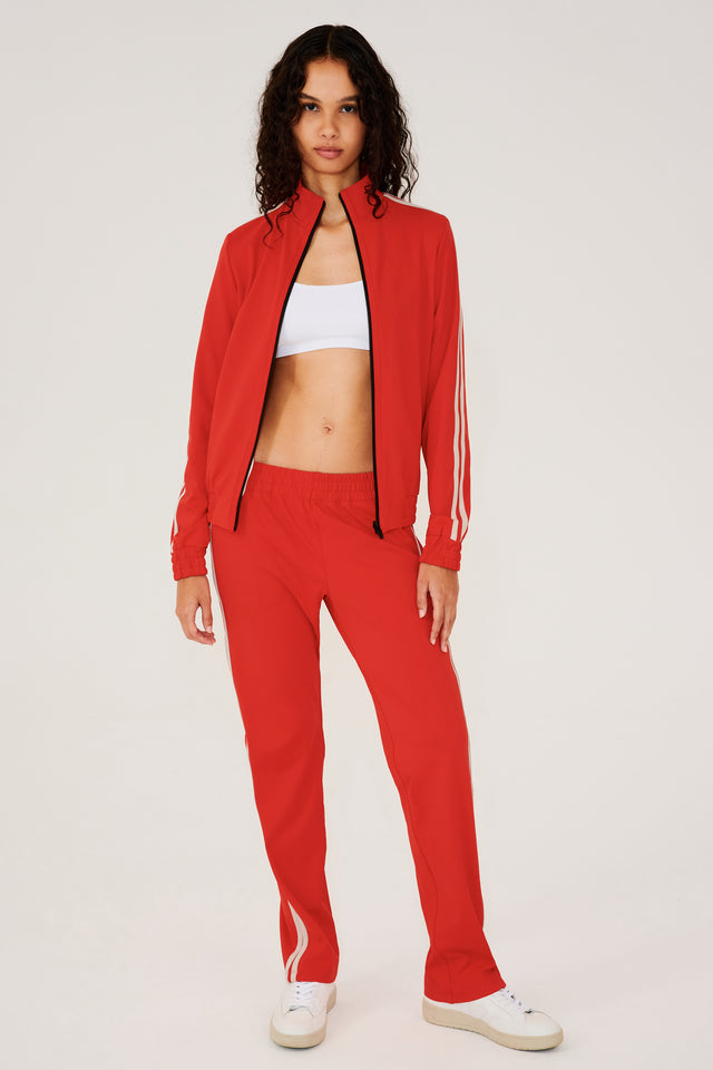 Full front view of girl wearing red zip jacket that stops under chin with two white stripes down the side and a black zipper in the front with a red sweatpants and white shoes