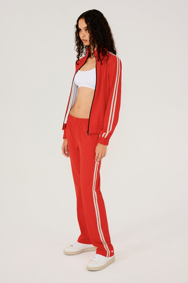Full side view of girl wearing red zip jacket that stops under chin with two white stripes down the side and a black zipper in the front with a red sweatpants and white shoes
