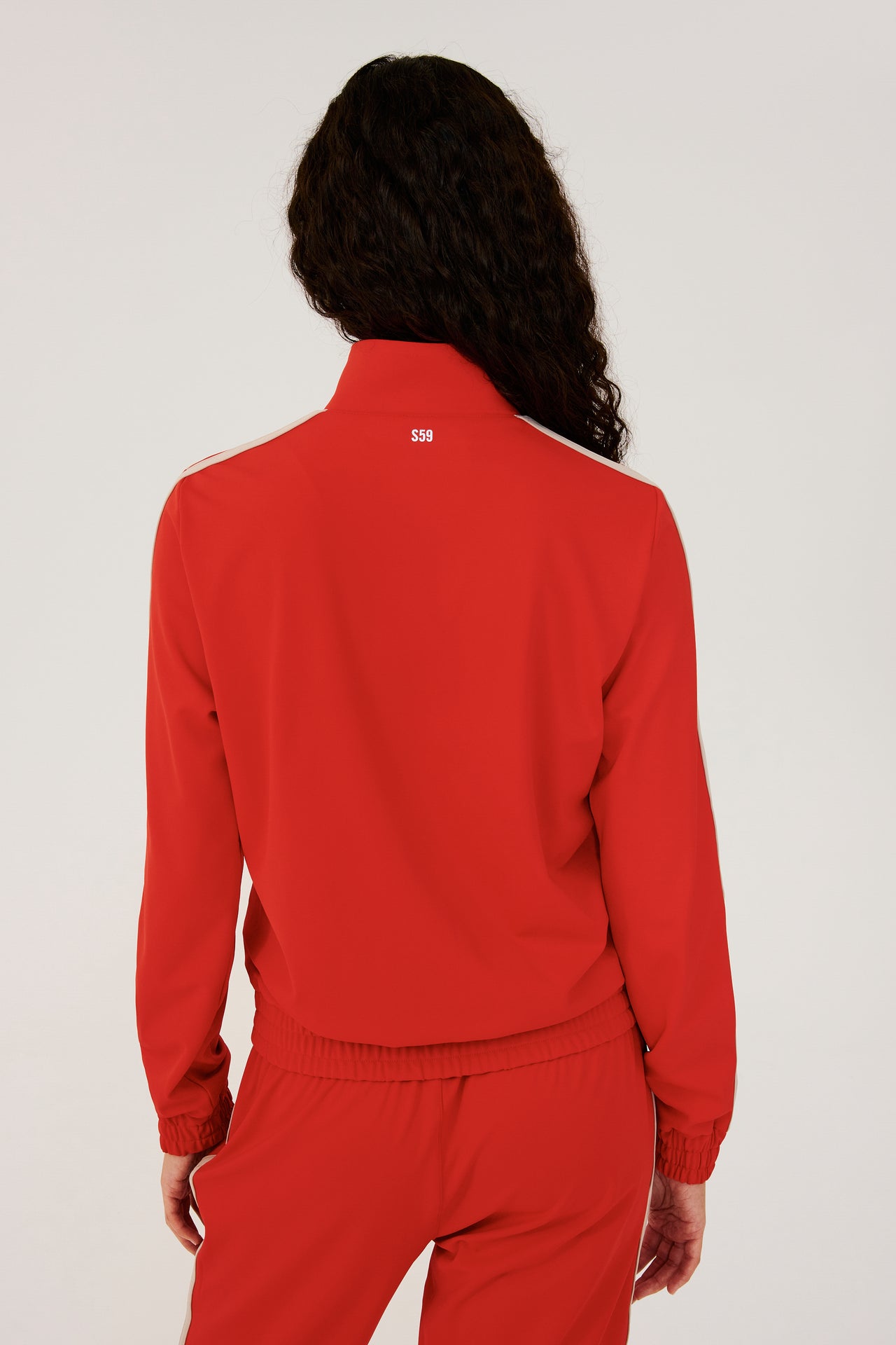 Back view of girl wearing red zip jacket that stops under chin with two white stripes down the side and a black zipper in the front with a red sweatpants