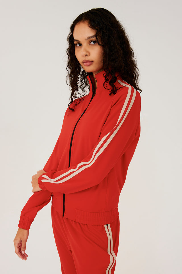 Side view of girl wearing red zip jacket that stops under chin with two white stripes down the side and a black zipper in the front with a red sweatpants 