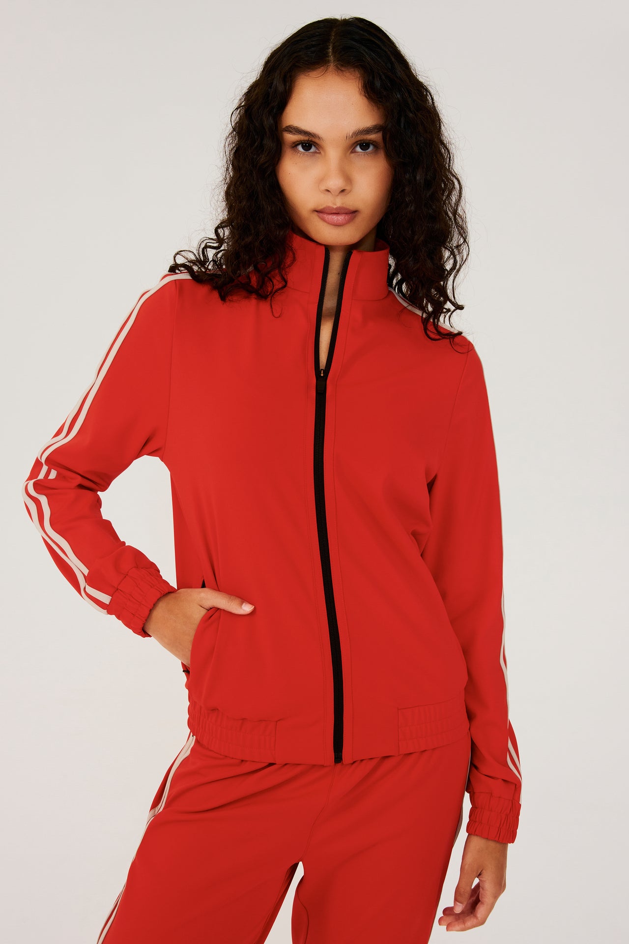 Front view of girl wearing red zip jacket that stops under chin with two white stripes down the side and a black zipper in the front with a red sweatpants