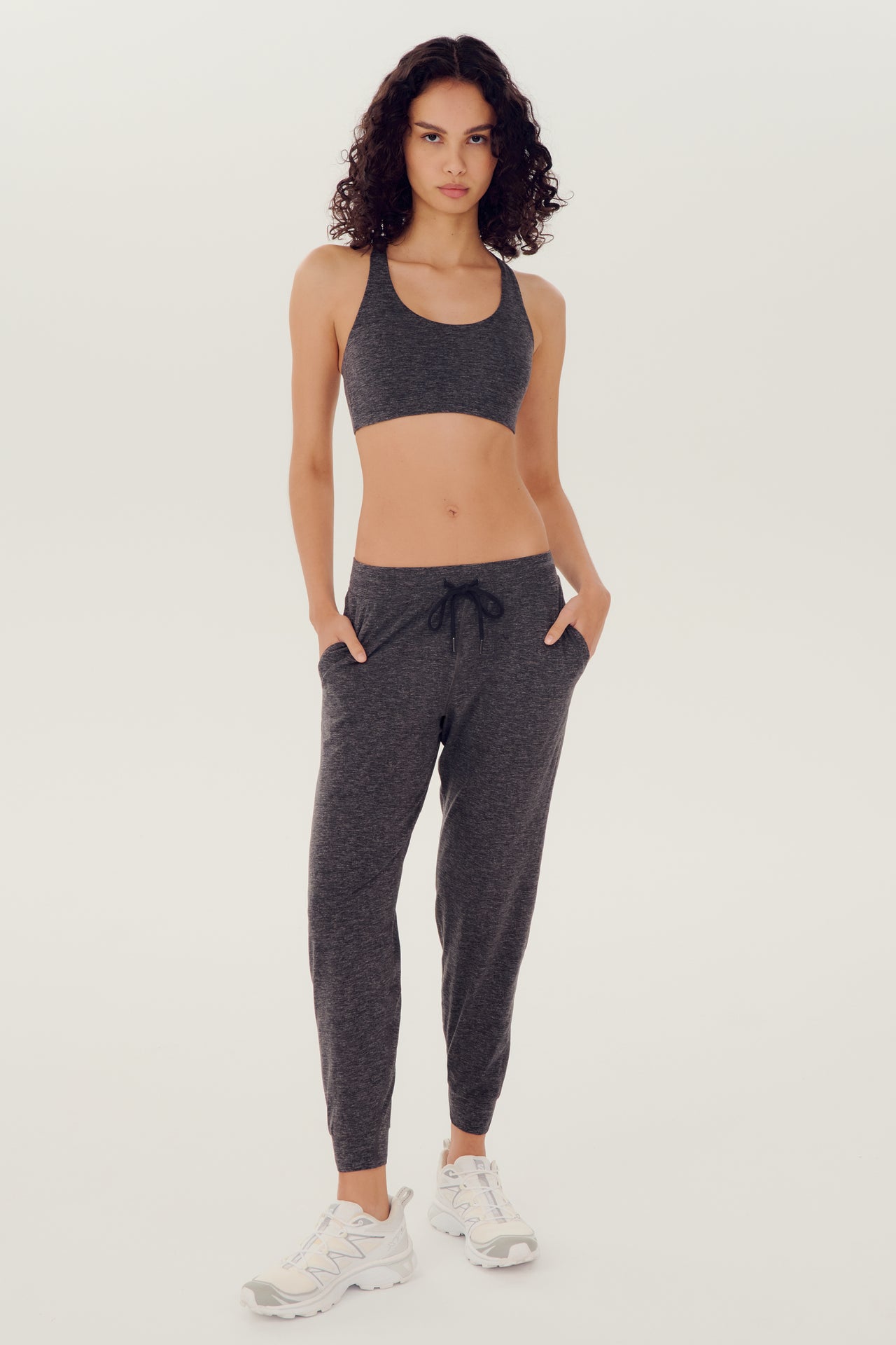 Full front view of girl wearing dark grey sweatpants with black tie around waistband and a dark grey sports bra with white shoes