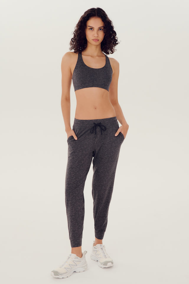 Full front view of girl wearing dark grey sweatpants with black tie around waistband and a dark grey sports bra with white shoes