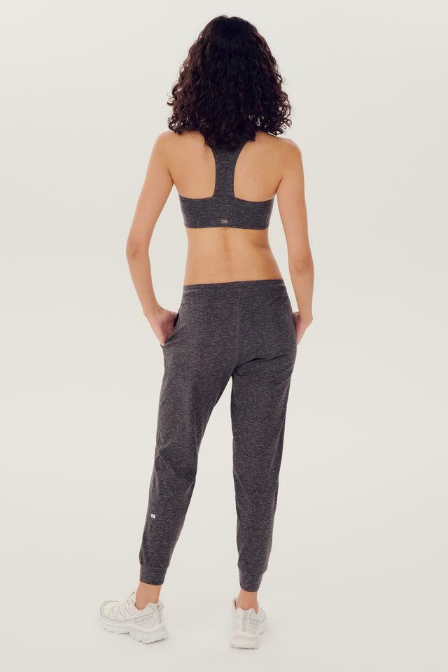 Full back view of girl wearing dark grey sweatpants with black tie around waistband and a dark grey sports bra with white shoes