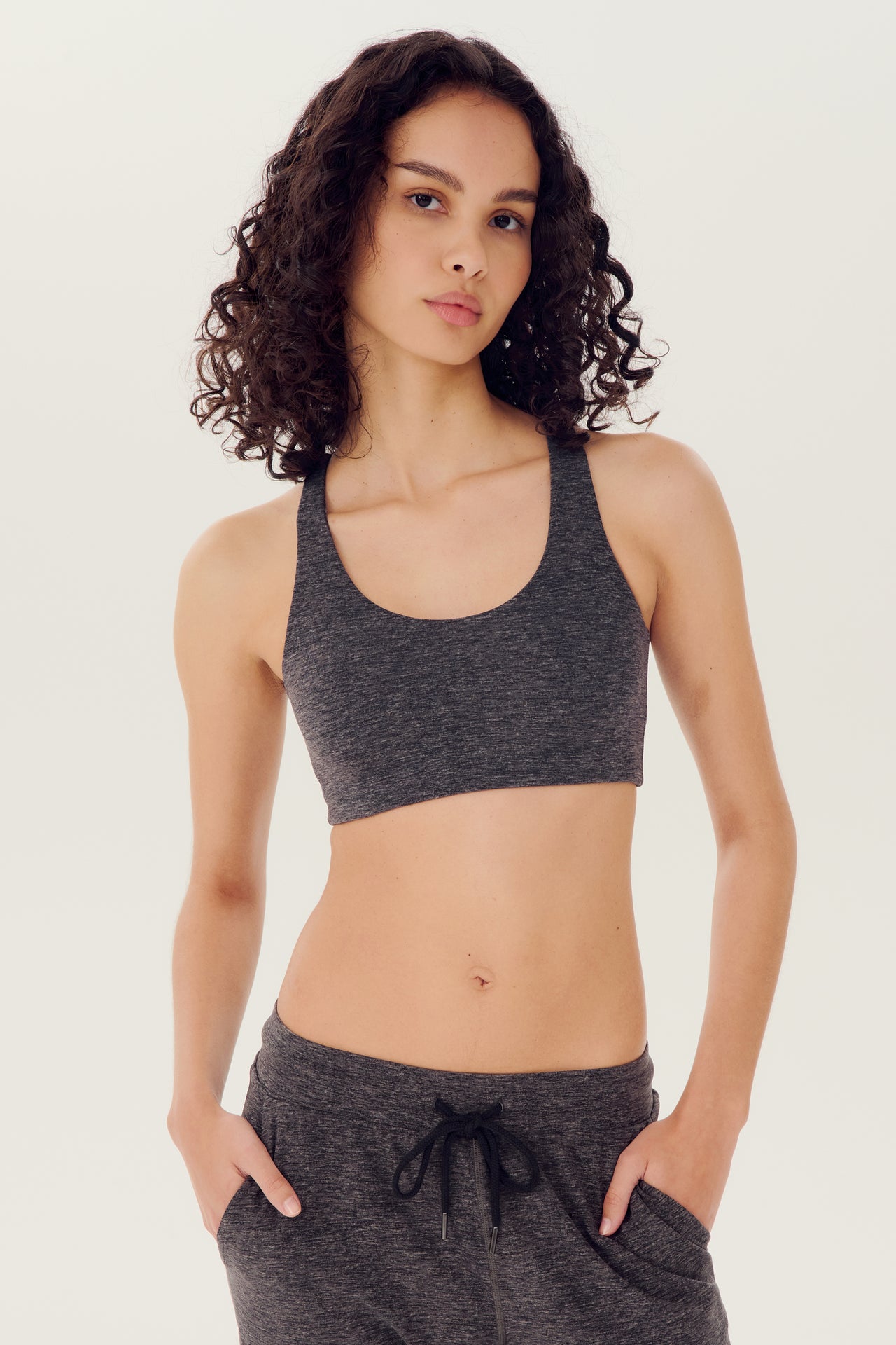 Front view of woman with dark wavy hair wearing dark grey bra and dark drey joggers with black drawstring