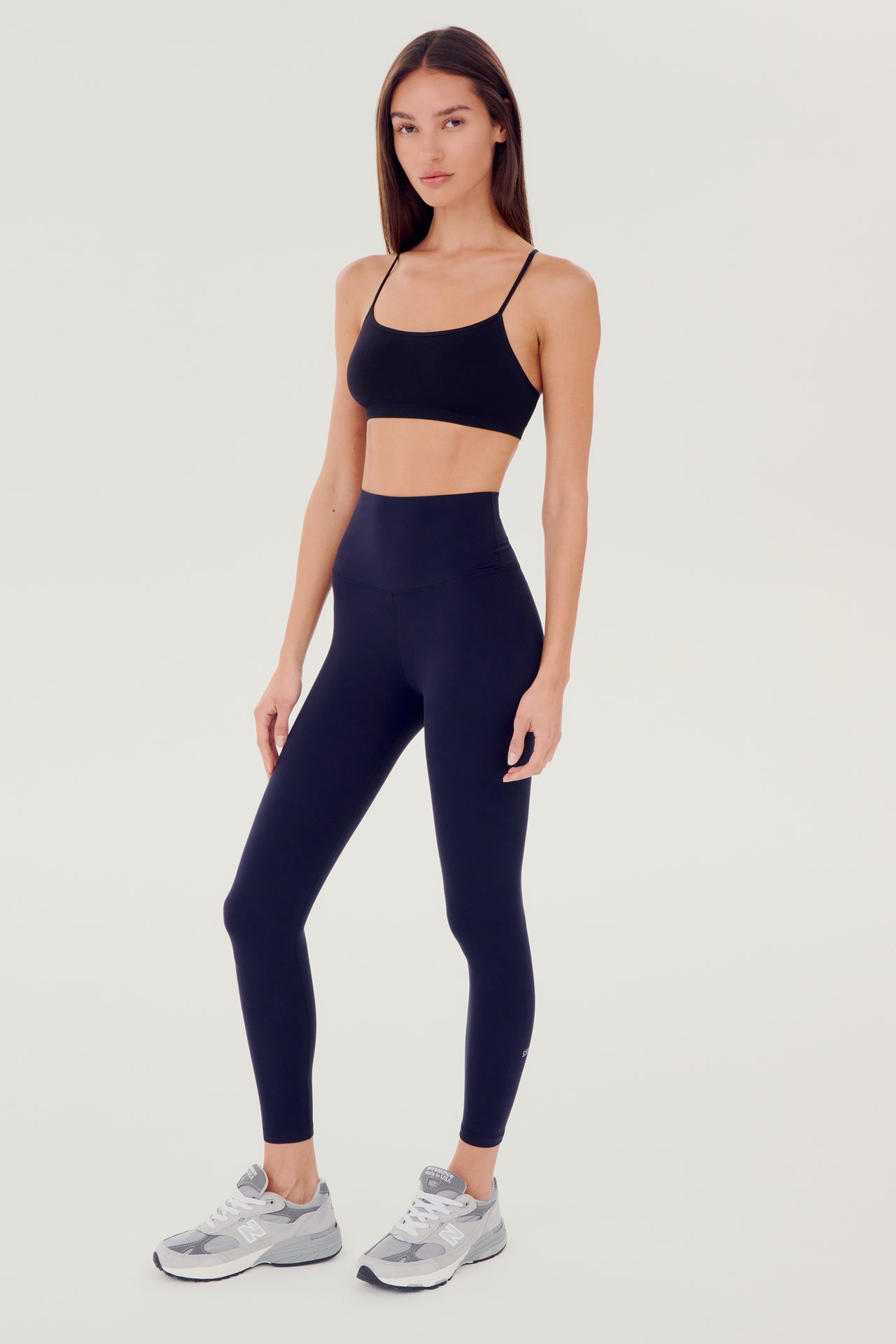 Full side view of girl wearing dark blue leggings right above the ankle with dark blue sports bra and grey shoes