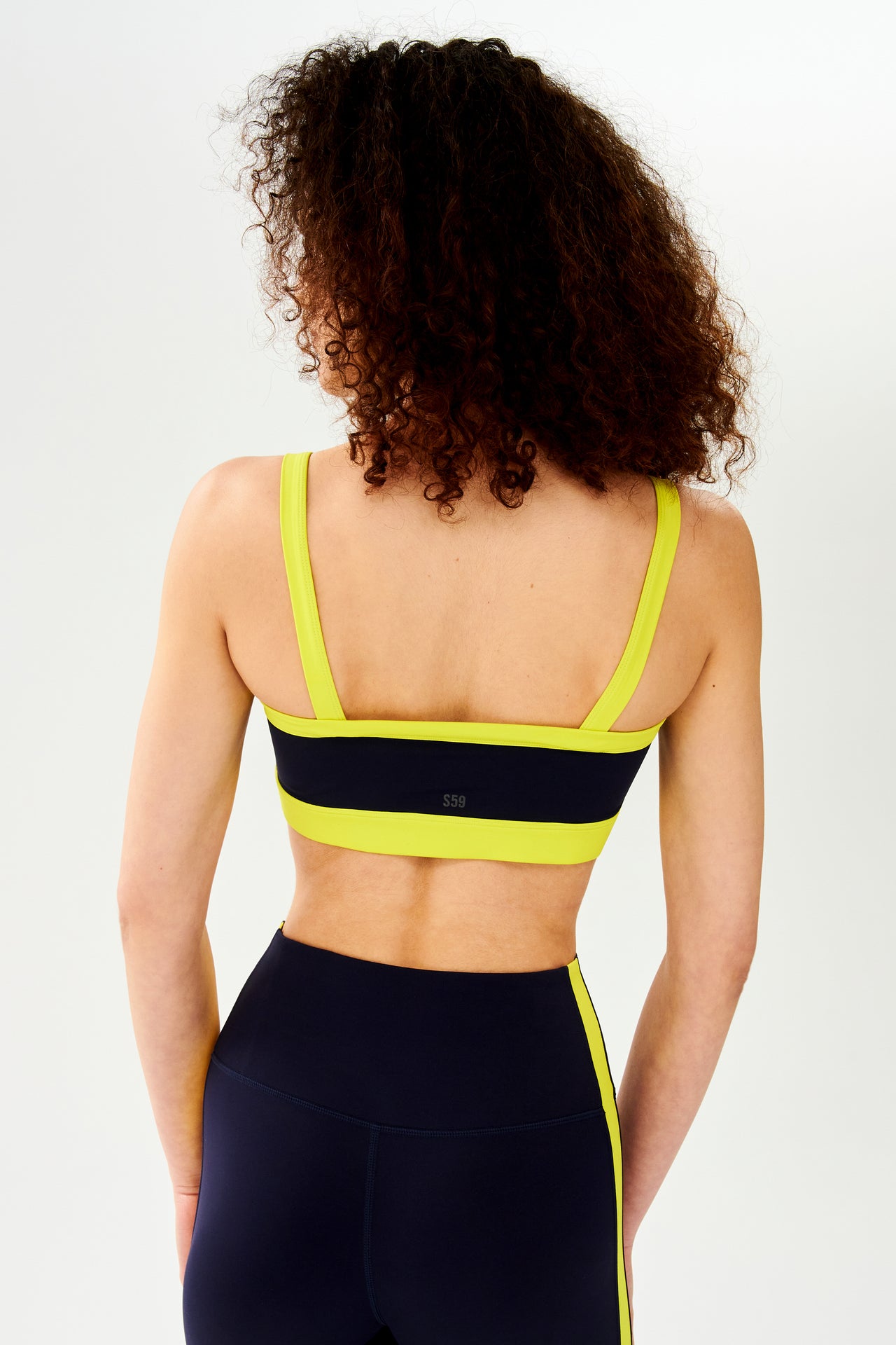 The back view of a woman wearing a SPLITS59 Monah Rigor Bra in Indigo/Chartreuse, designed for maximum workout comfort and support.