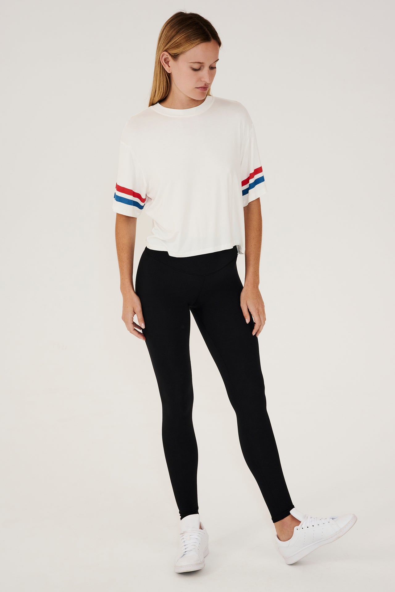 Full front view of girl wearing a short sleeve cropped white shirt with red and blue stripes on the arm and a black leggings with white shoes