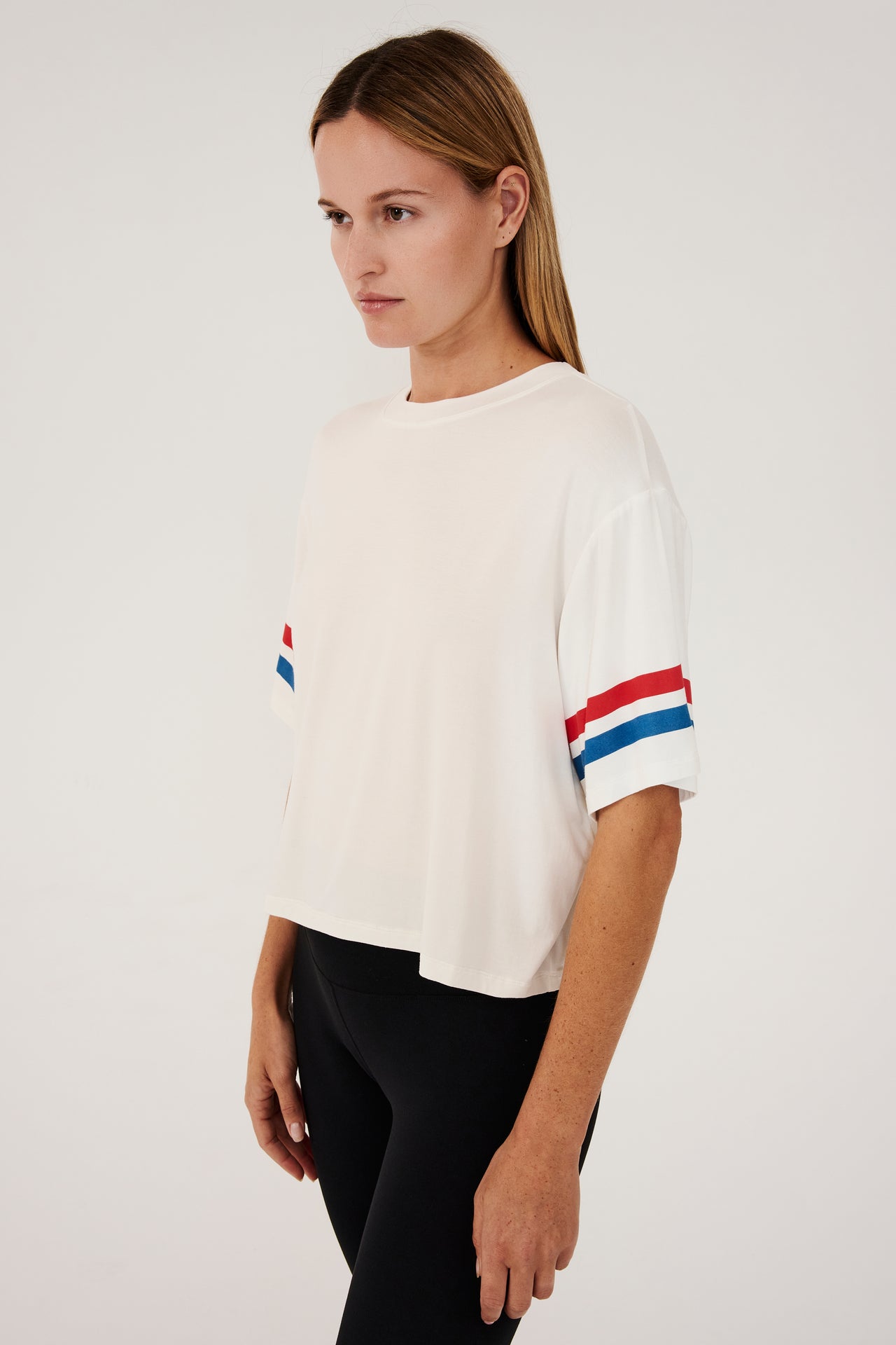 Side view of girl wearing a short sleeve cropped white shirt with red and blue stripes on the arm and a black leggings 