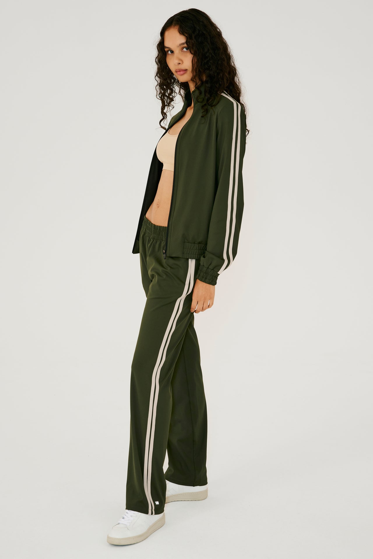 Full side view of girl wearing dark green zip jacket that stops under chin with two white stripes down the side and a dark green sweatpants with white shoes