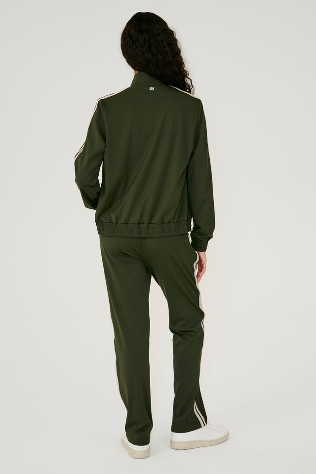 Full back view of girl wearing dark green zip jacket that stops under chin with two white stripes down the side and a dark green sweatpants with white shoes