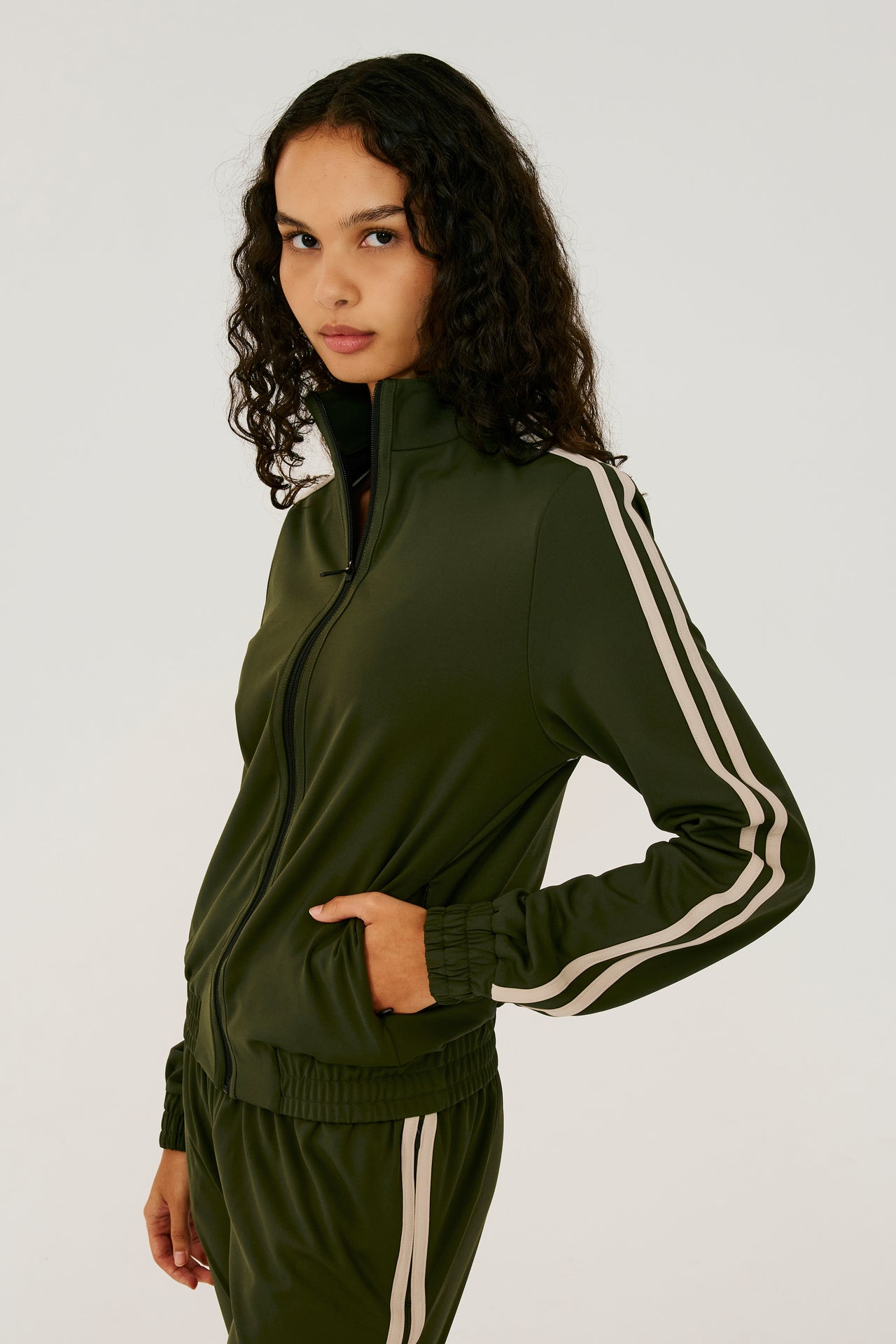Side view of girl wearing dark green zip jacket that stops under chin with two white stripes down the side and a dark green sweatpants