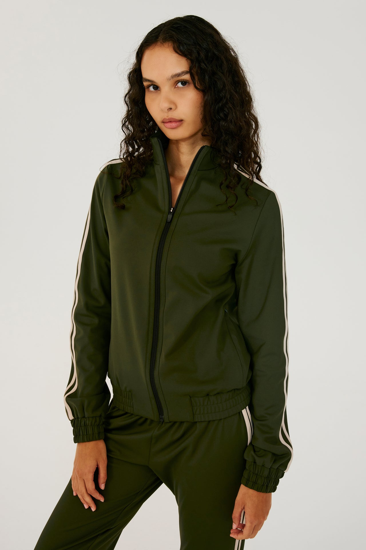 Front view of girl wearing dark green zip jacket that stops under chin with two white stripes down the side and a dark green sweatpants
