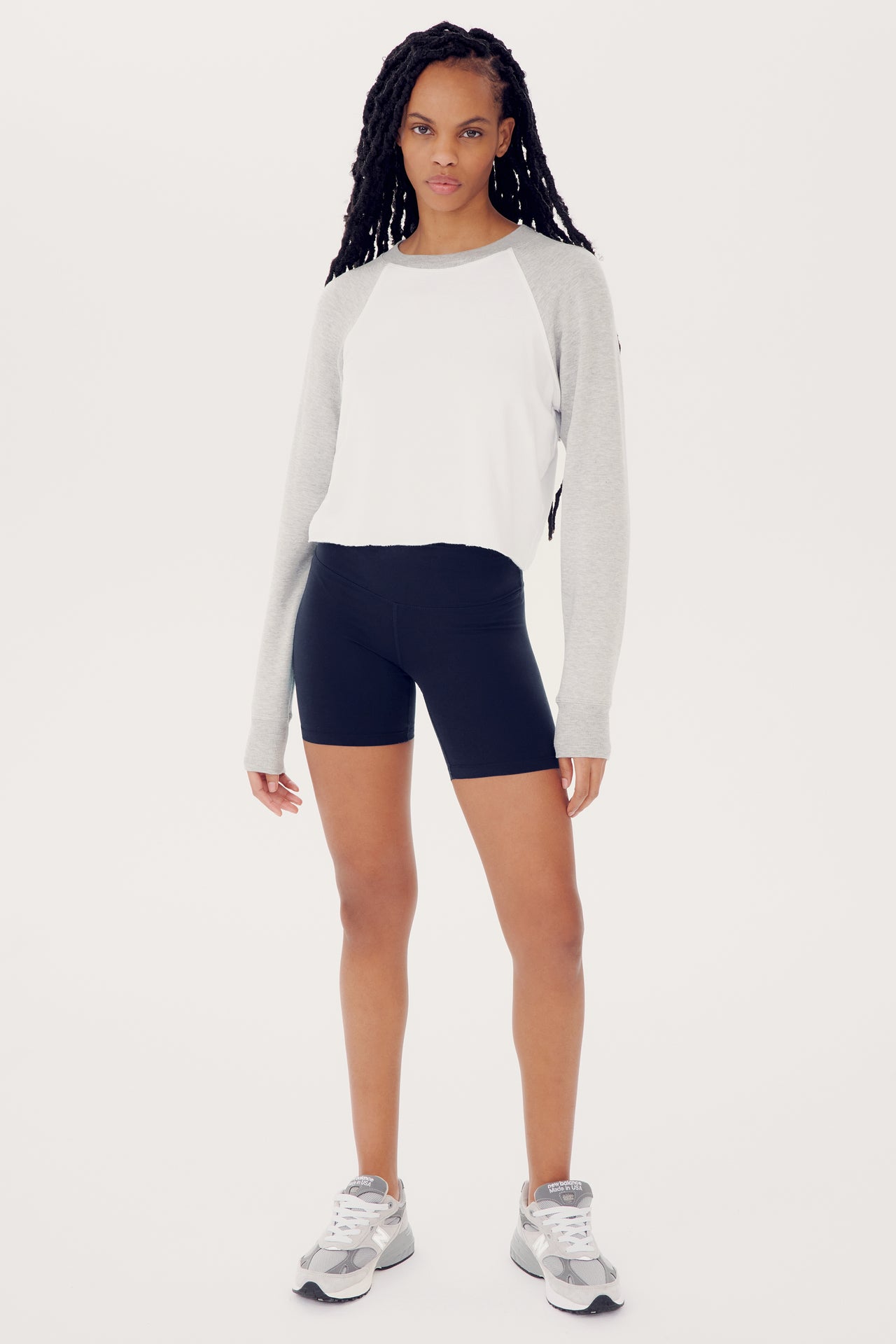 A woman in a Warm Up Crop Fleece Sweatshirt by SPLITS59 made of comfort fabric and black spandex shorts standing confidently, viewed against a plain white backdrop.