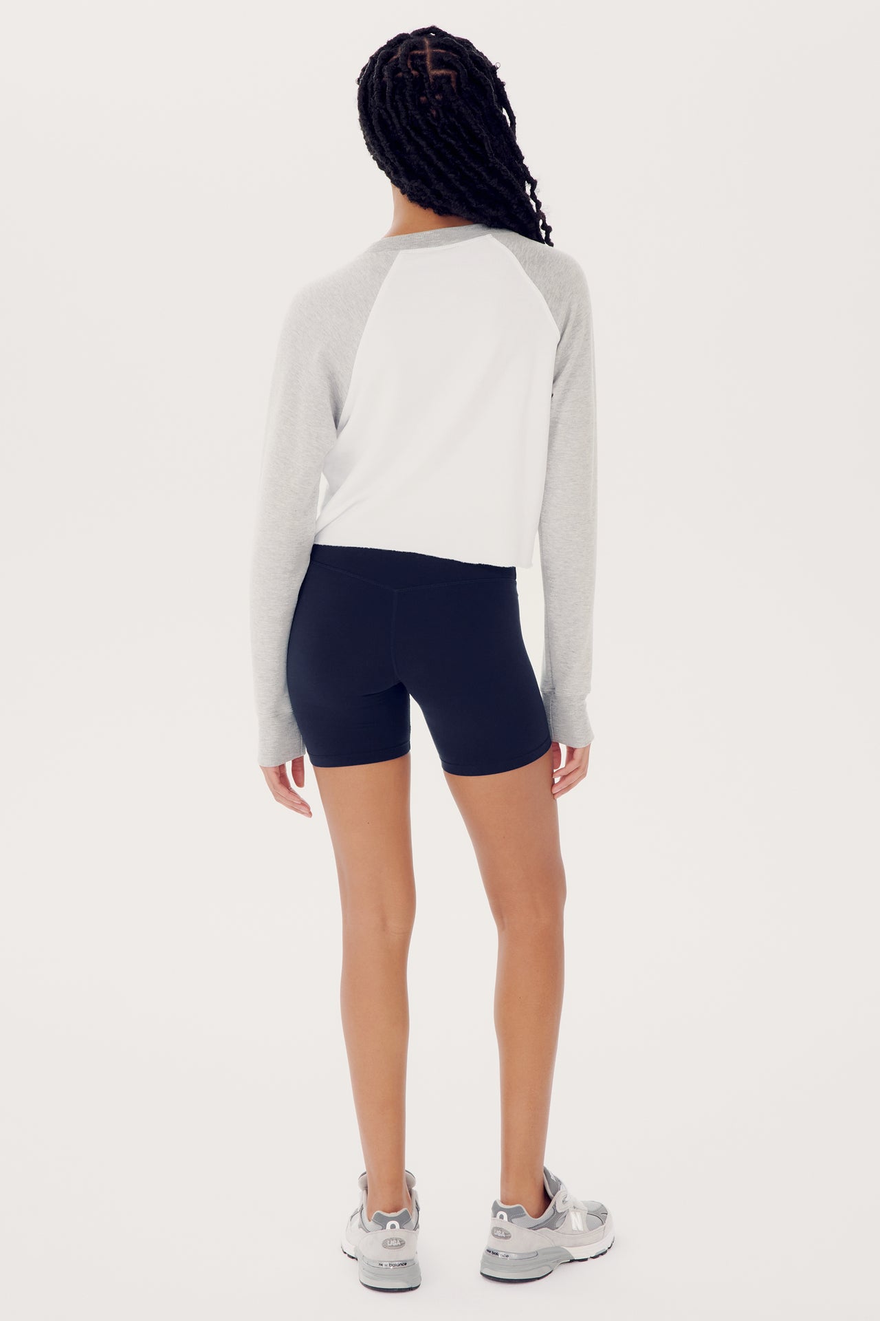 A woman standing with her back to the camera, wearing a SPLITS59 Warm Up Crop Fleece Sweatshirt in Heather Grey/White and navy blue spandex shorts, on a white background.