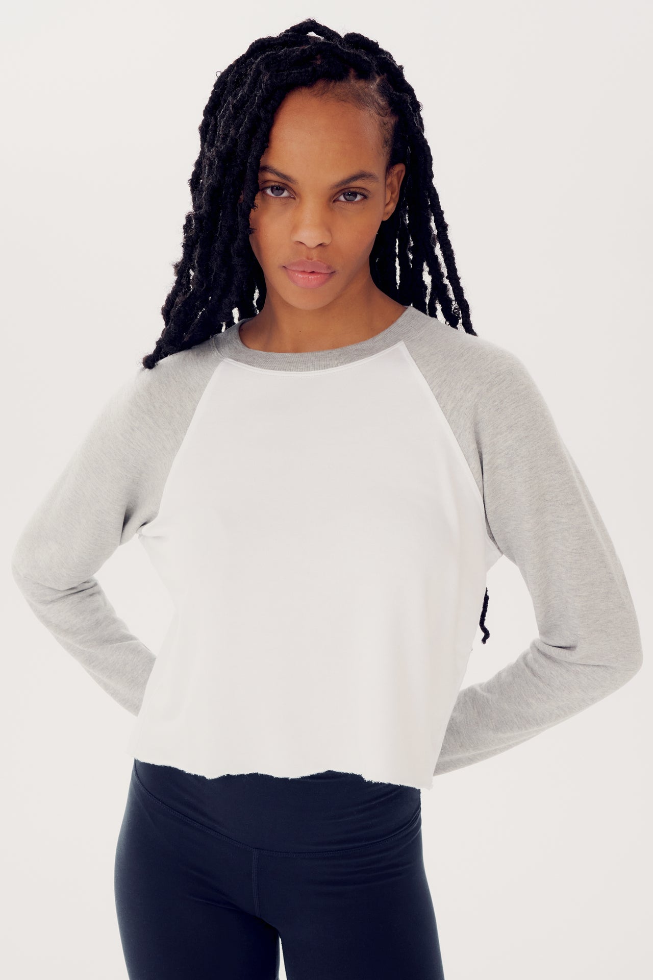 A woman with long braids wearing a SPLITS59 Warm Up Crop Fleece Sweatshirt in Heather Grey/White and black leggings, standing with hands on hips and looking at the camera.