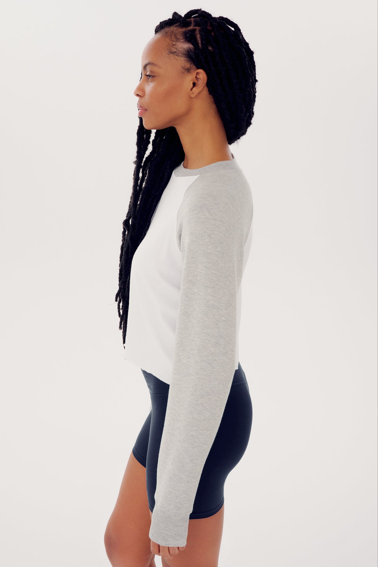 Woman in a SPLITS59 Warm Up Crop Fleece Sweatshirt in Heather Grey/White and navy spandex shorts, standing sideways with long braided hair.