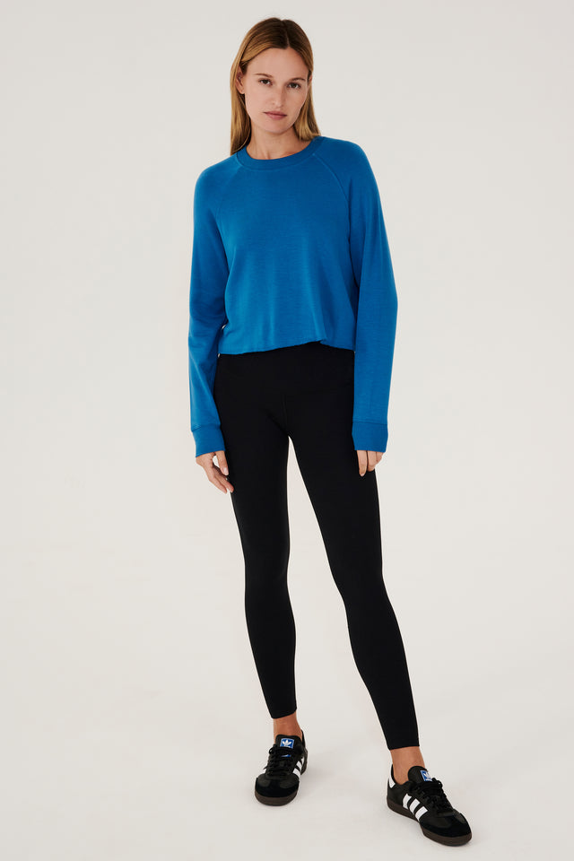 Full front view of woman with dark blonde hair, wearing cropped  bright blue sweatshirt with black leggings and black shoes with white stripes