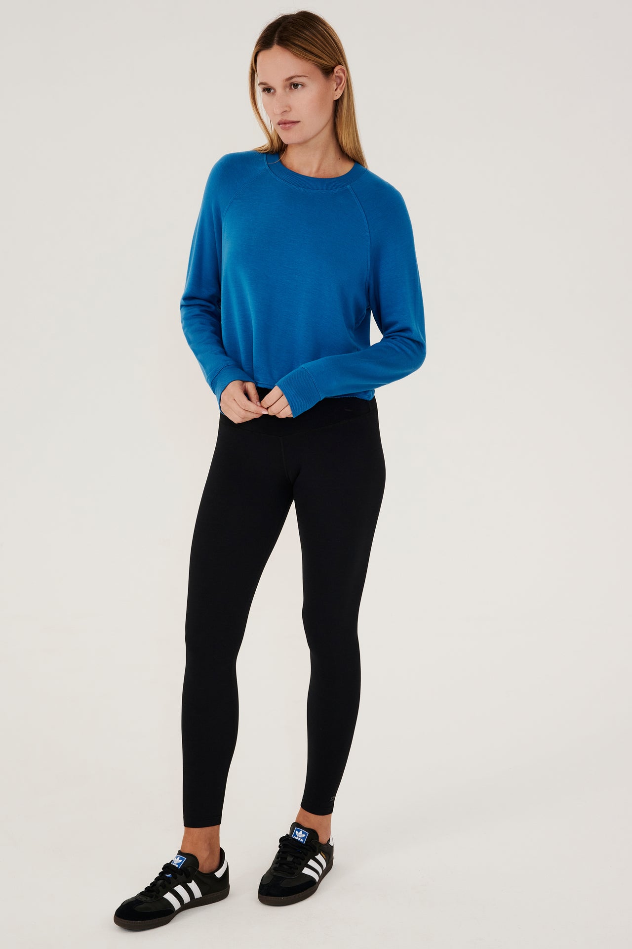 Full front view of woman with dark blonde hair, wearing cropped  bright blue sweatshirt with black leggings and black shoes with white stripes