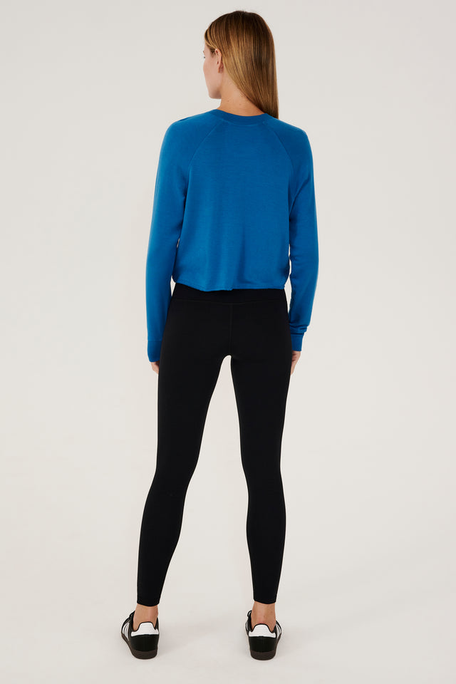 Full back view of woman with dark blonde hair, wearing cropped  bright blue sweatshirt with black leggings and black shoes with white stripes