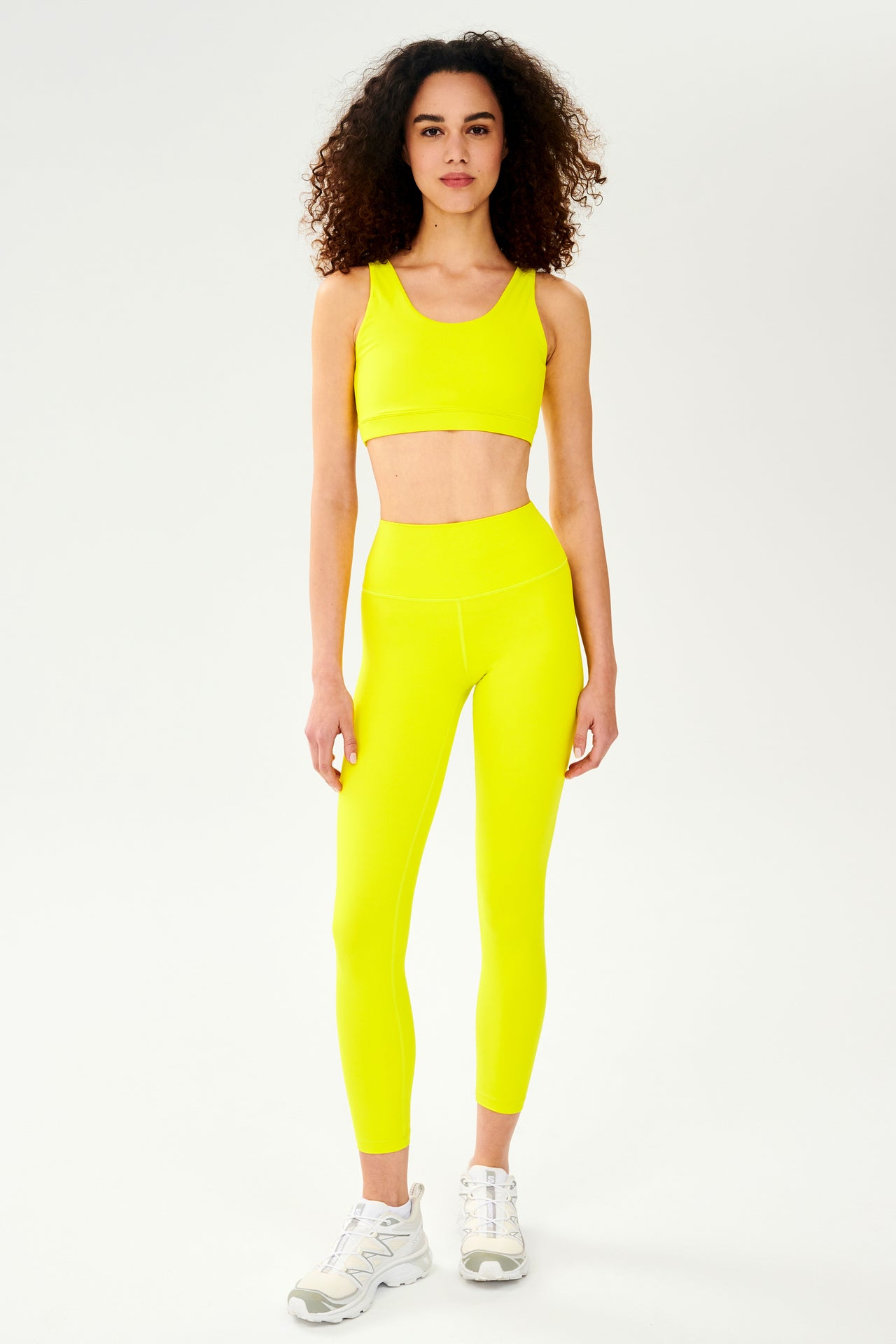 Front full view of woman with curly dark wearing bright yellow bra with bright yellow high waist  leggings and white shoes