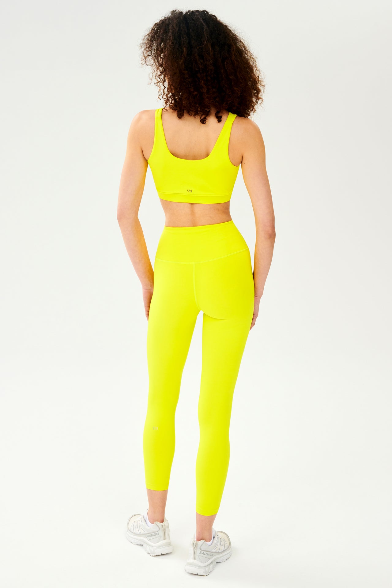 Back full view view of woman with curly dark wearing bright yellow bra with bright yellow high waist  leggings and white shoes