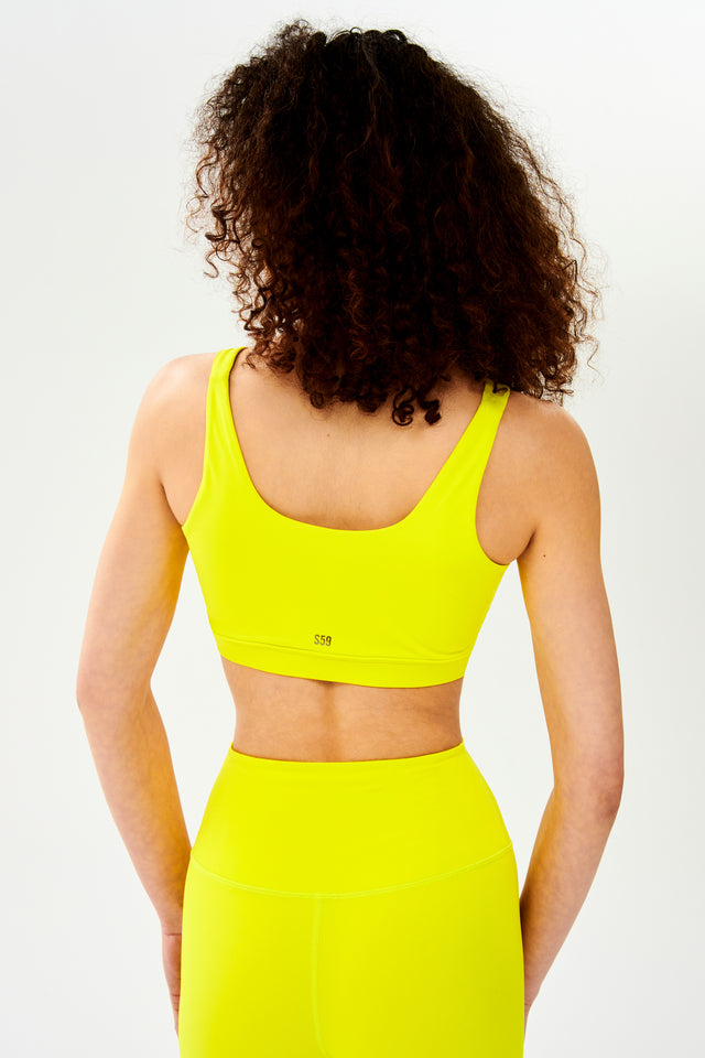 Back  view of woman with curly dark wearing bright yellow bra with black S59 logo and bright yellow leggings 