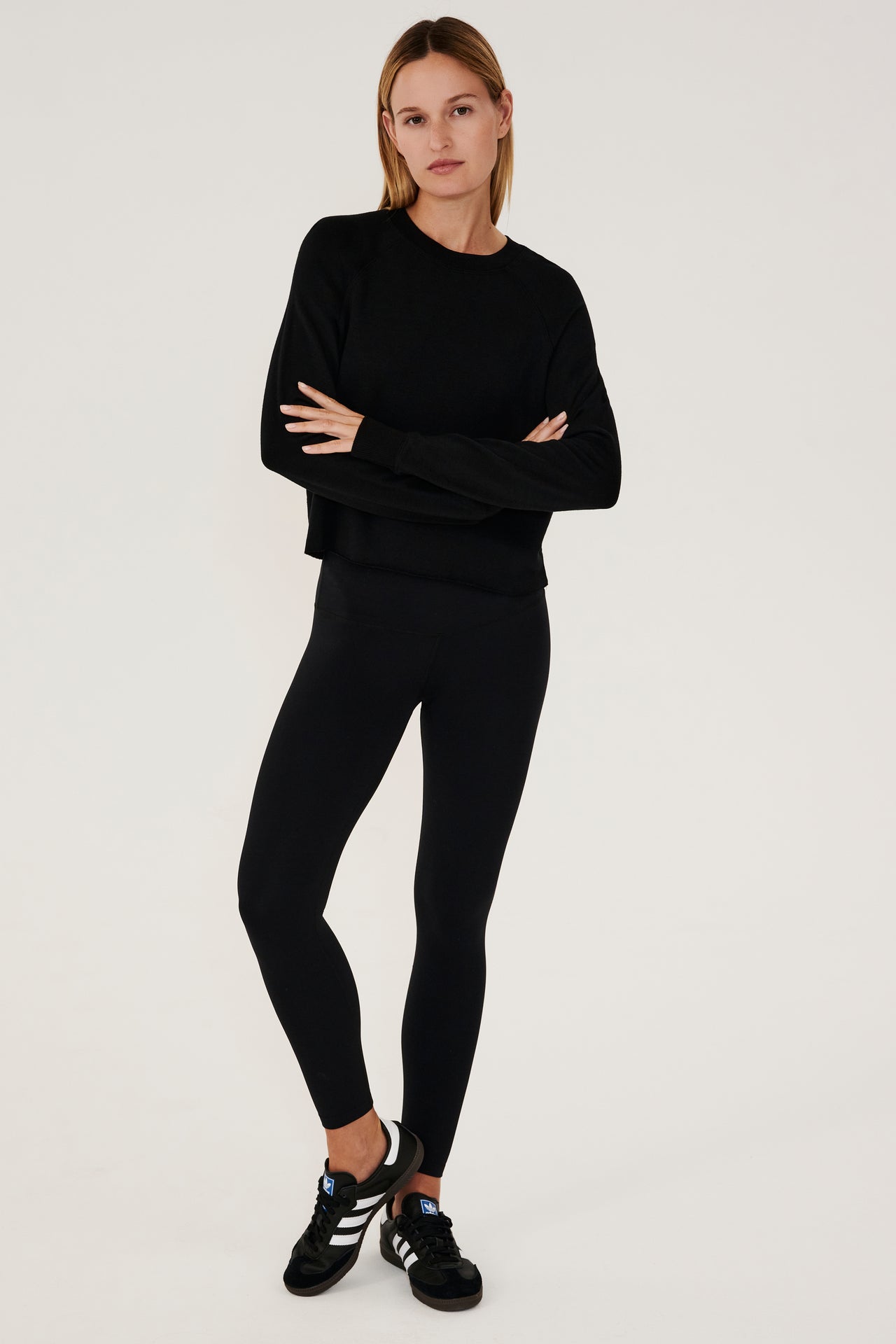 Full front view of woman with dark blonde hair, wearing black cropped sweatshirt with black leggings and wearing black shoes with white stripes