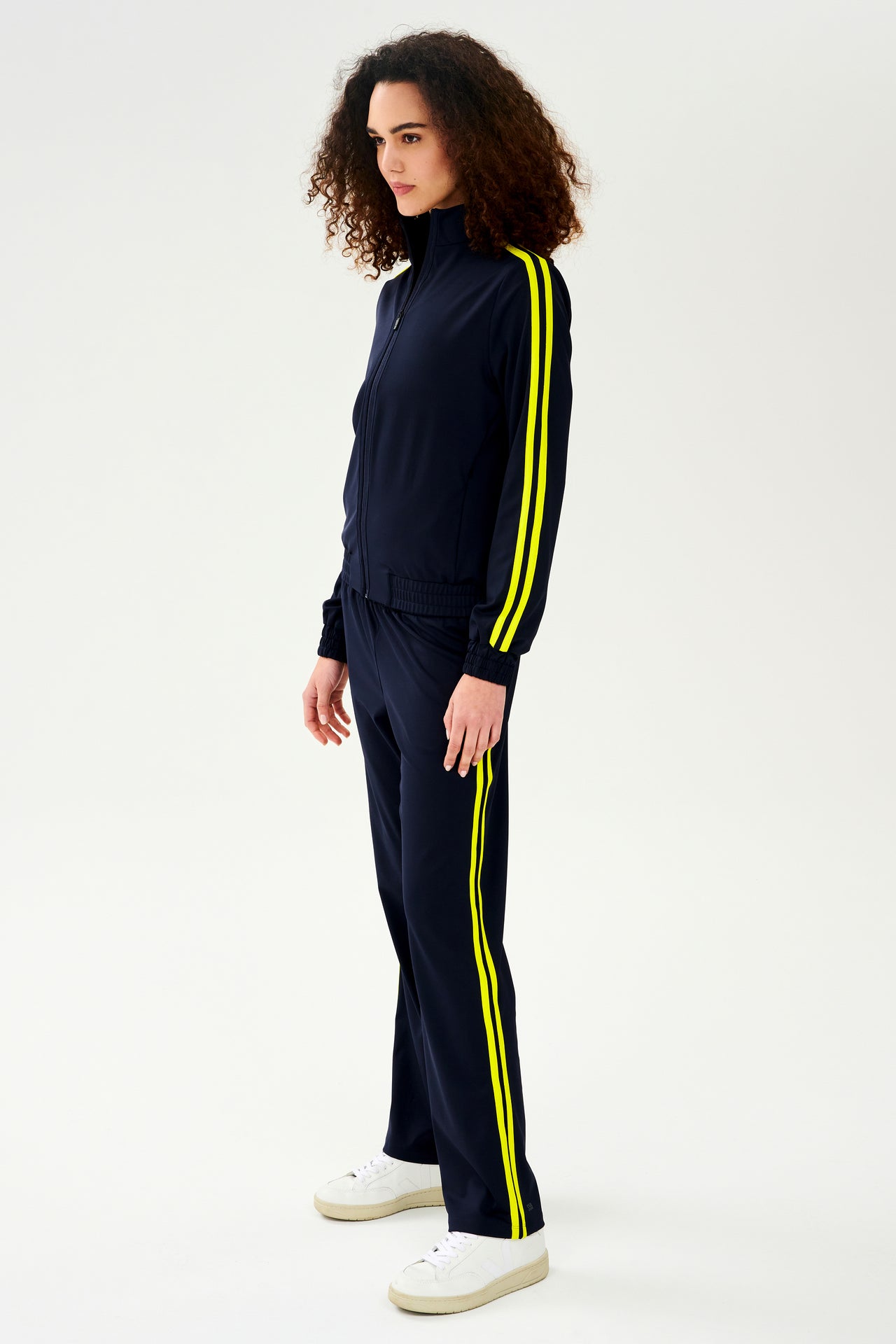 A woman wearing a luxurious SPLITS59 Fox Techflex Jacket in Indigo/Chartreuse with yellow side stripes, perfect as an après workout piece.