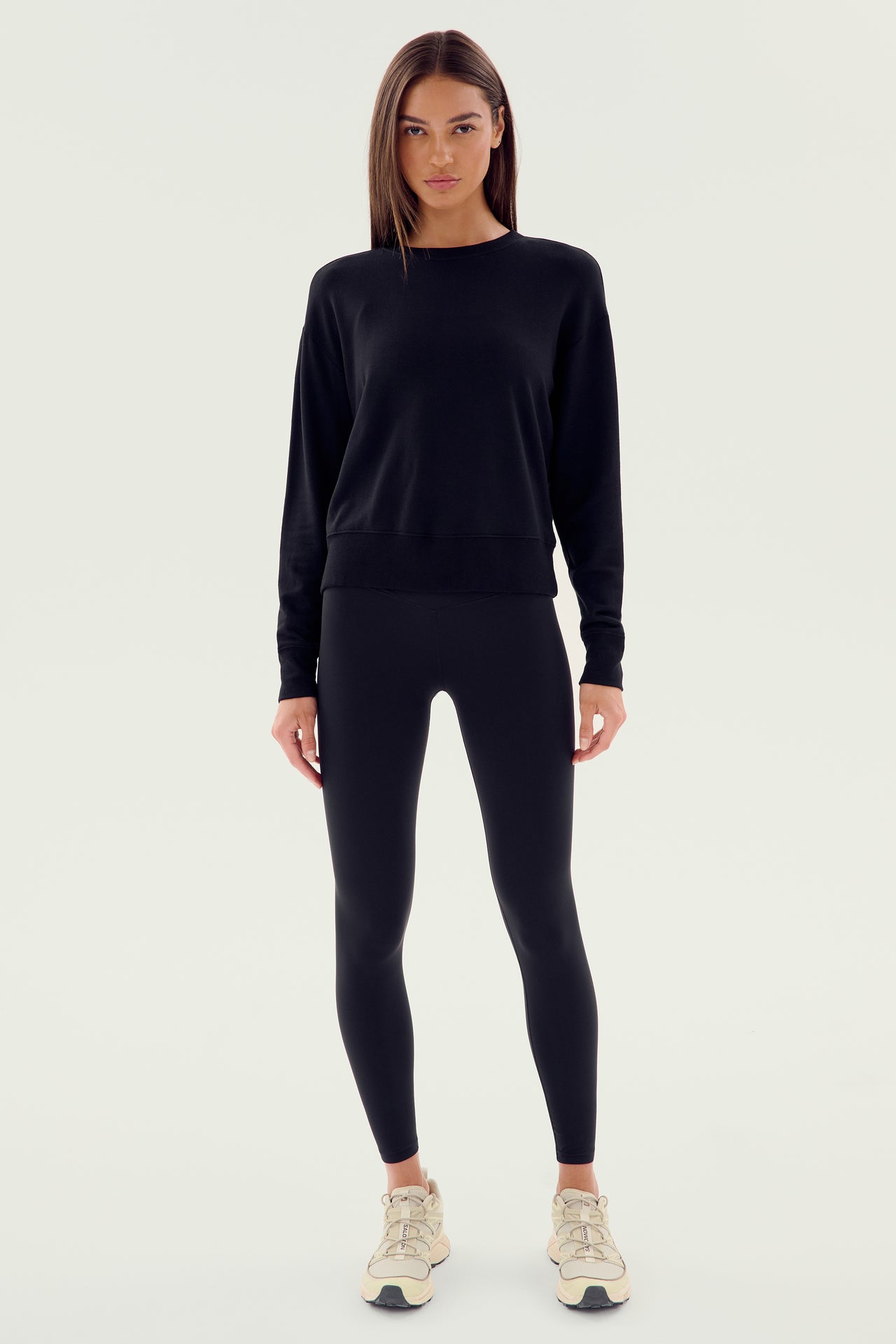 A person standing against a plain background, wearing the SPLITS59 Sonja Fleece Sweatshirt in black, black leggings, and light-colored athletic shoes.