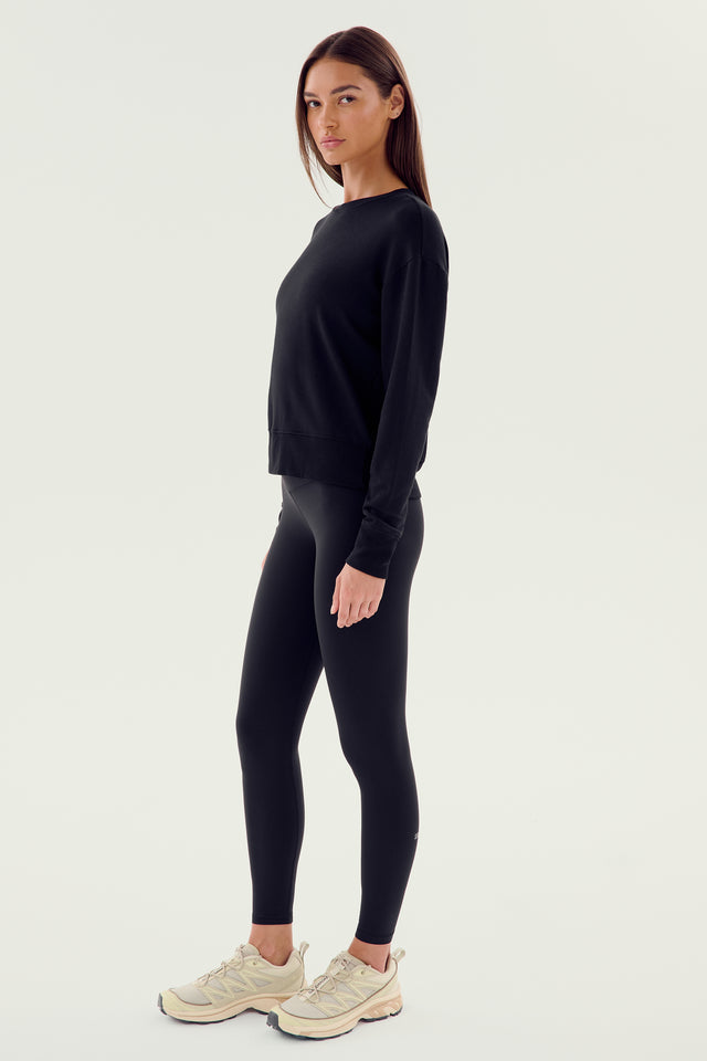 A woman stands against a plain background, wearing the SPLITS59 Sonja Fleece Sweatshirt in ultra luxe fleece, accompanied by black leggings and white sneakers, facing slightly to the left.