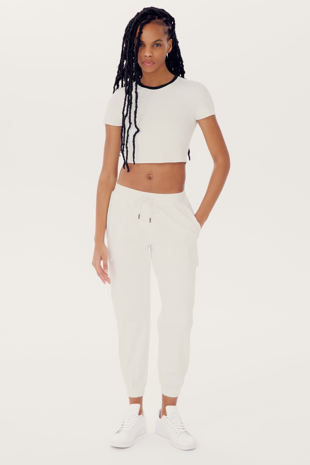 A person with long braided hair is wearing a white crop top and relaxed fit, mid-weight fabric SPLITS59 Supplex Cargo Pant - White, paired with white sneakers, standing against a plain white background.