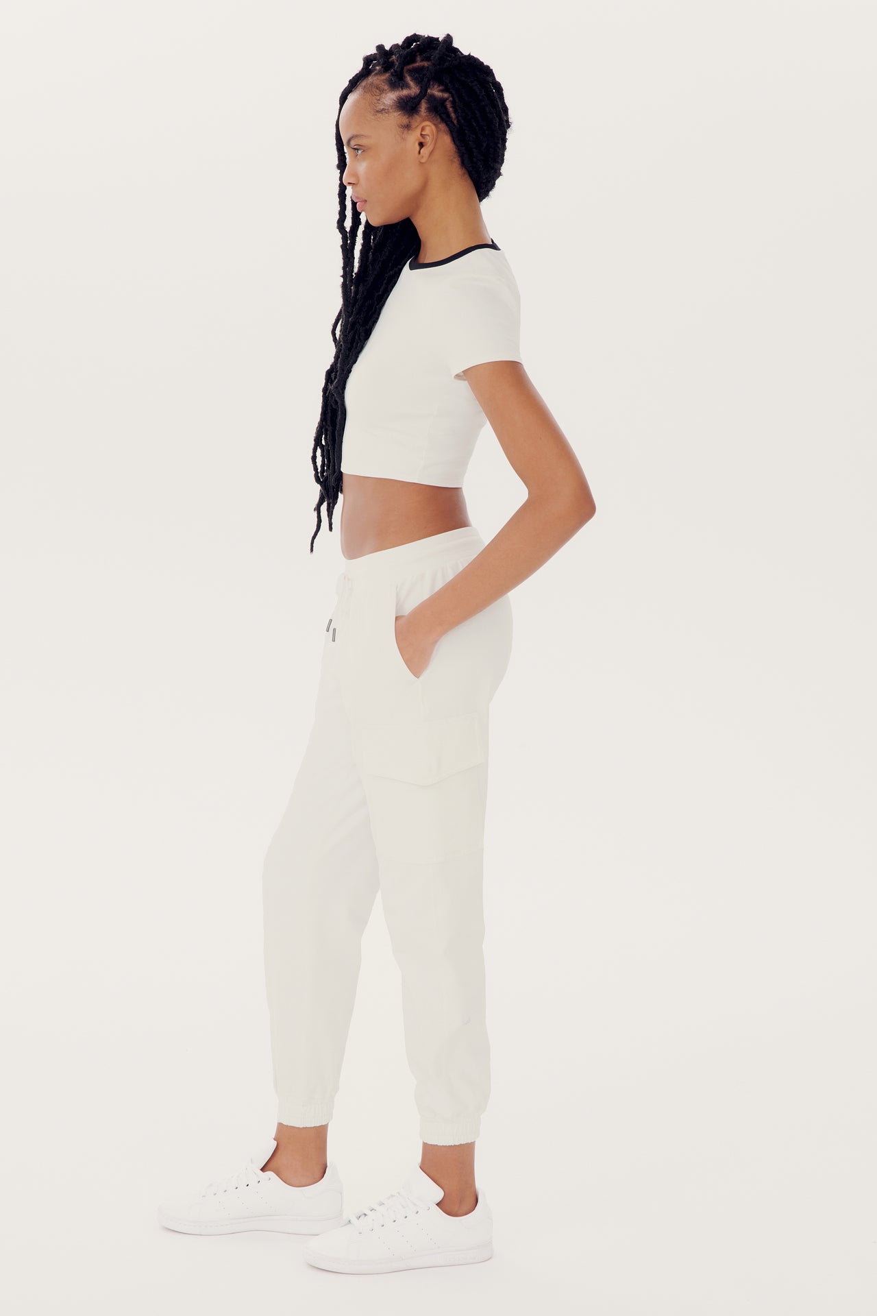 A person with braided hair is wearing a white crop top, Supplex Cargo Pant - White by SPLITS59, and white sneakers, standing in a side profile view against a plain white background.