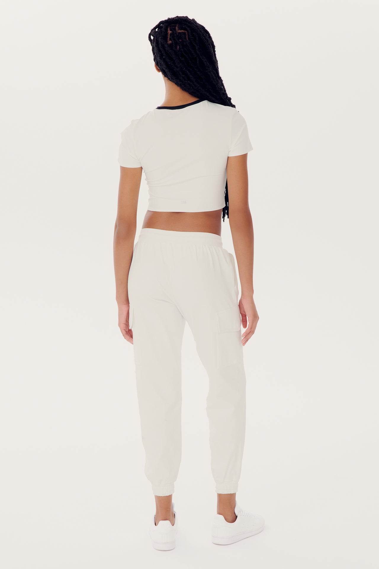 A person with long braided hair stands facing away from the camera, wearing a short-sleeved crop top and relaxed fit SPLITS59 Supplex Cargo Pant - White in mid-weight fabric, all paired with white sneakers.
