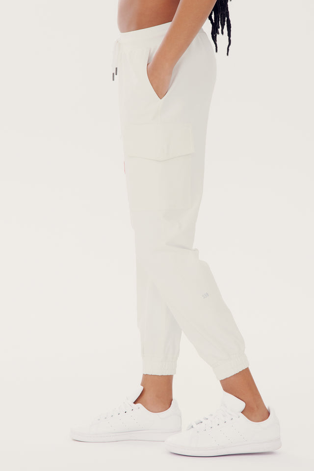 A person is shown from the side wearing relaxed fit, light-colored Supplex Cargo Pant - White by SPLITS59 with elastic cuffs and white sneakers. The person has one hand in their pocket.