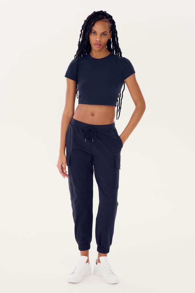 A woman standing in a neutral pose wearing a black SPLITS59 Airweight S/S Crop - Indigo designed for gym workouts, dark jogger pants, and white sneakers.